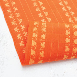 100% cotton fabric in a vibrant stunning orange color with a cream woven textured floral motif arranged in vertical rows on a white background.