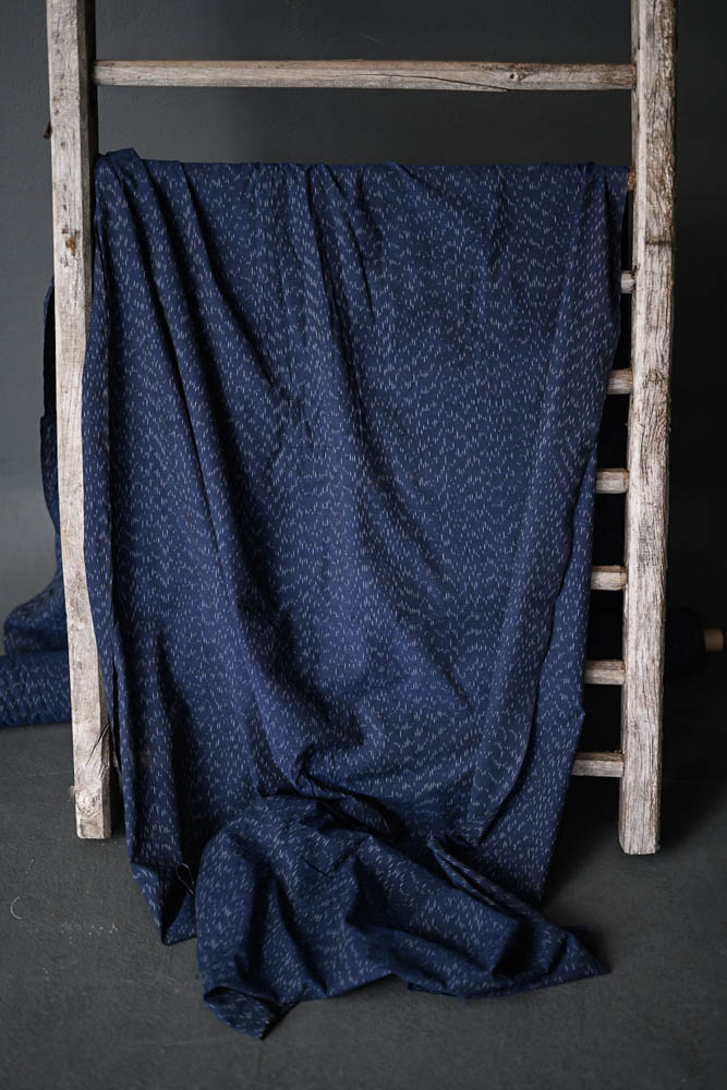 Indigo Snow Cotton Ikat fabric draped over a ladder on a dark grey backdrop 