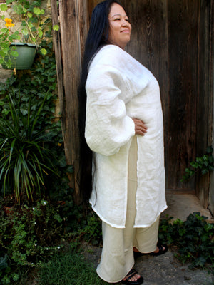 A woman with long black hair stands outdoors in front of a rustic wooden door, wearing a loose white long-sleeve tunic layered over cream wide-leg pants and black sandals.