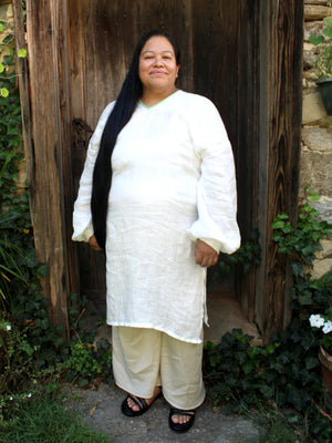 A woman with long black hair stands in front of a rustic wooden door wearing a white long-sleeve tunic and light beige pants, with black sandals. Green foliage and a stone wall are visible around the doorway.