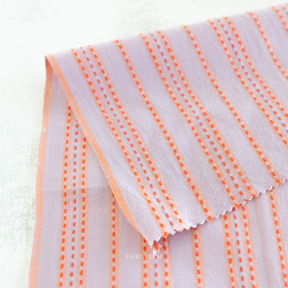 Close up of lavender woven cotton with an orange stitched stripe on a white background.