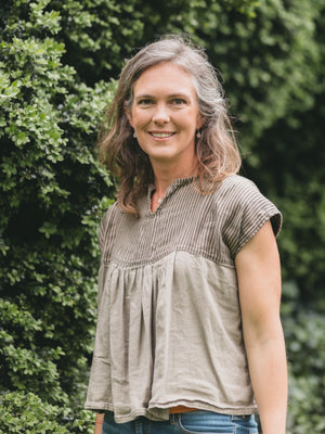 A woman with shoulder-length gray-brown hair stands outdoors in front of a leafy green hedge, wearing a loose taupe-beige short-sleeve top with a smocked yoke and blue jeans.