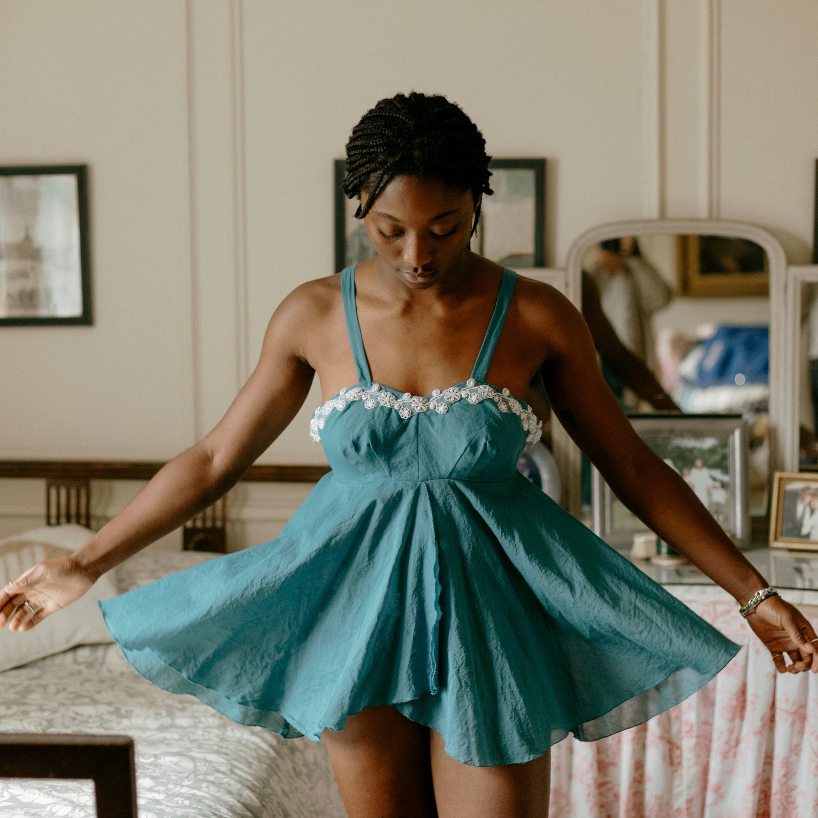 Black woman twirling so her skirt billows out, wearing a teal colored short lingerie dress with white lace trim at the top. In a bedroom. 