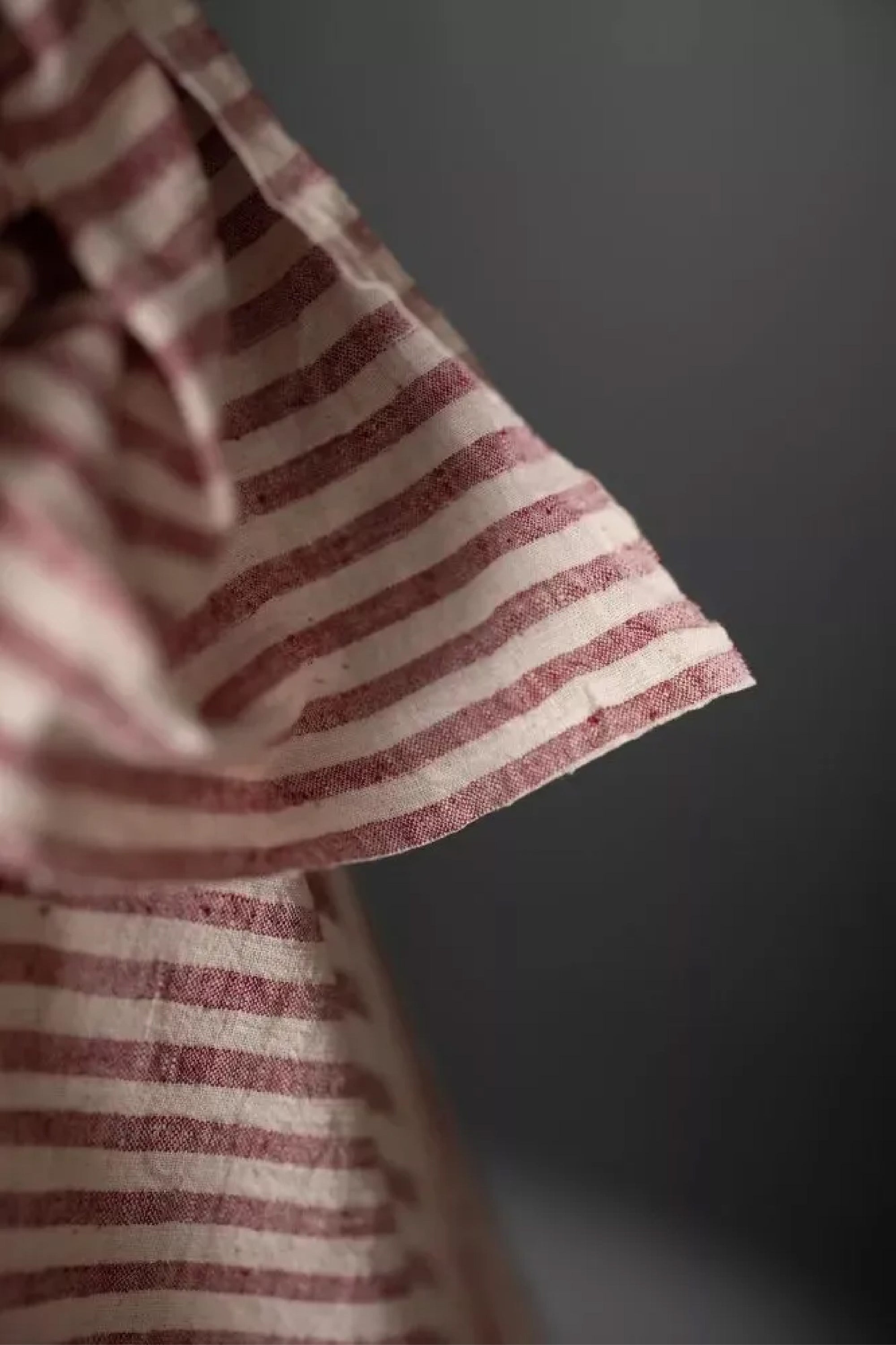 close up of a handloomed candy stripe of dusty red and natural with a loose weave and a washed out slubby texture fabric on a dark grey background.