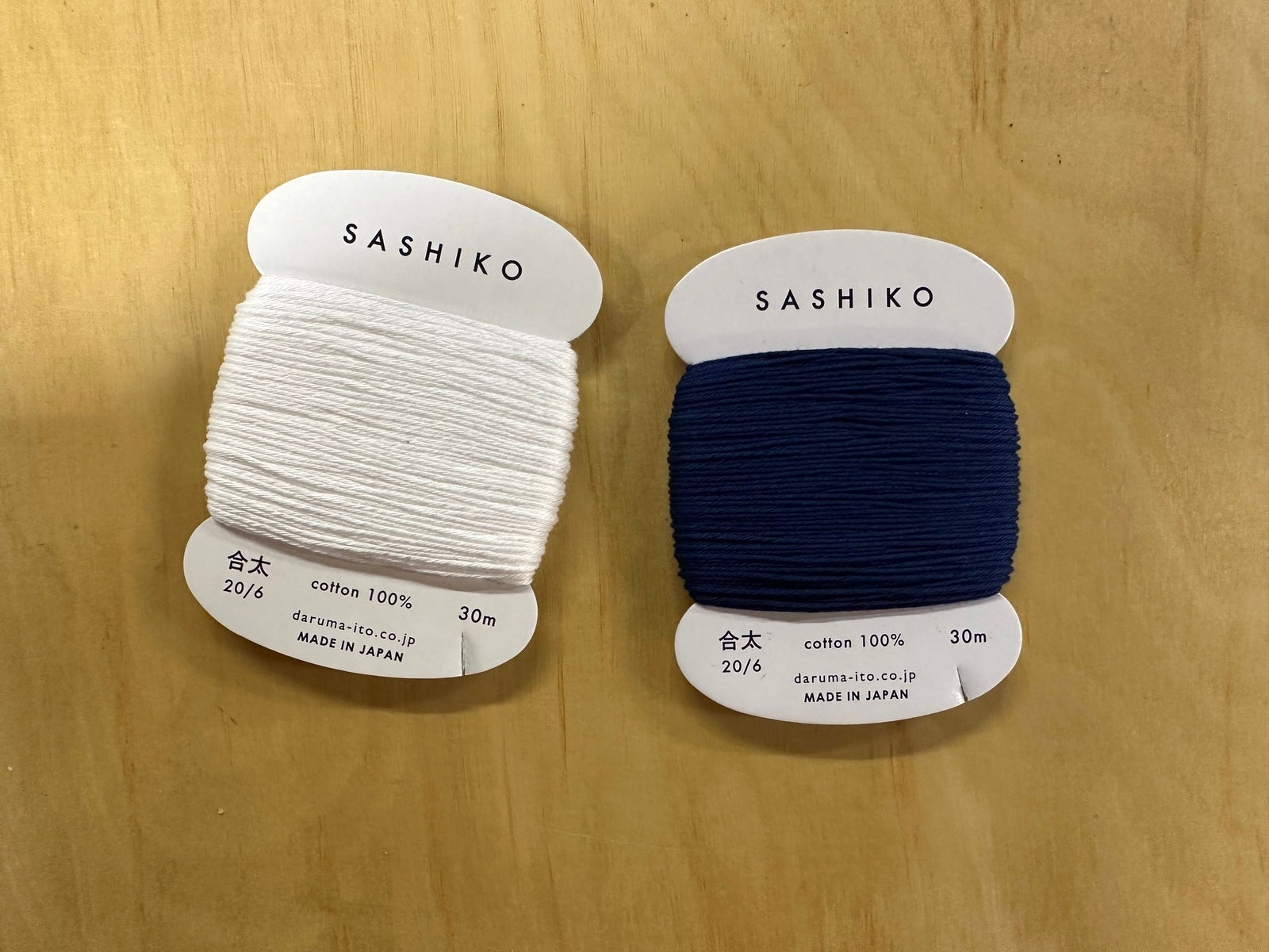 Two spools of Sashiko thread, one white and one navy, on a wooden surface.