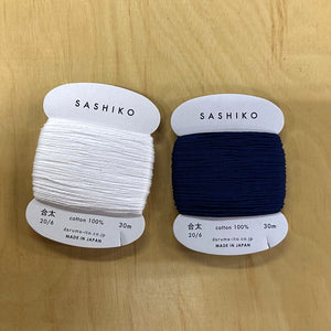 Two spools of Sashiko thread, one white and one navy, on a wooden surface.