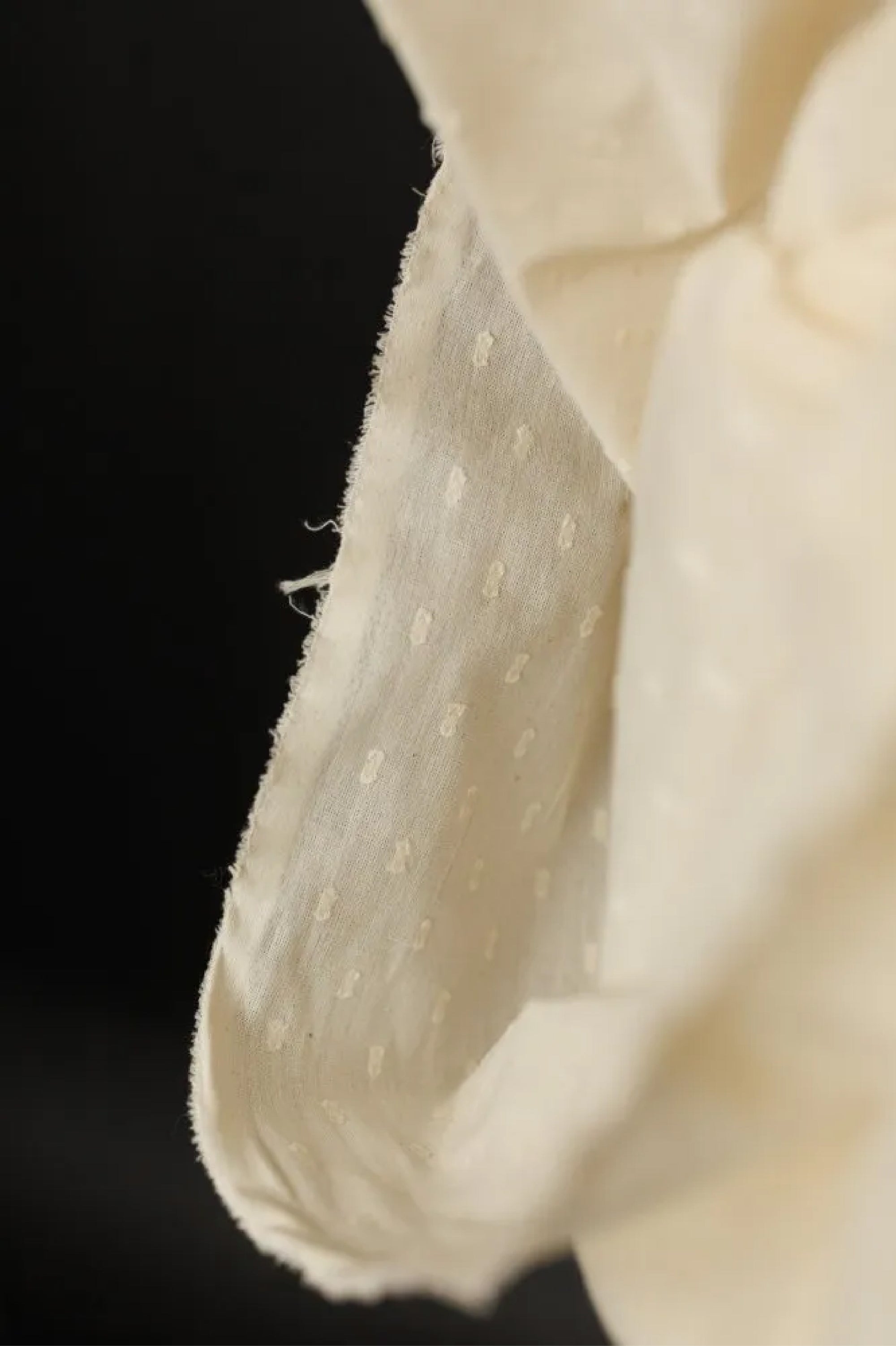 Close up of a natural Indian lightweight cotton in  a off-white color with a swiss dot pattern repeating ever 2.5cm. on a very dark grey background.