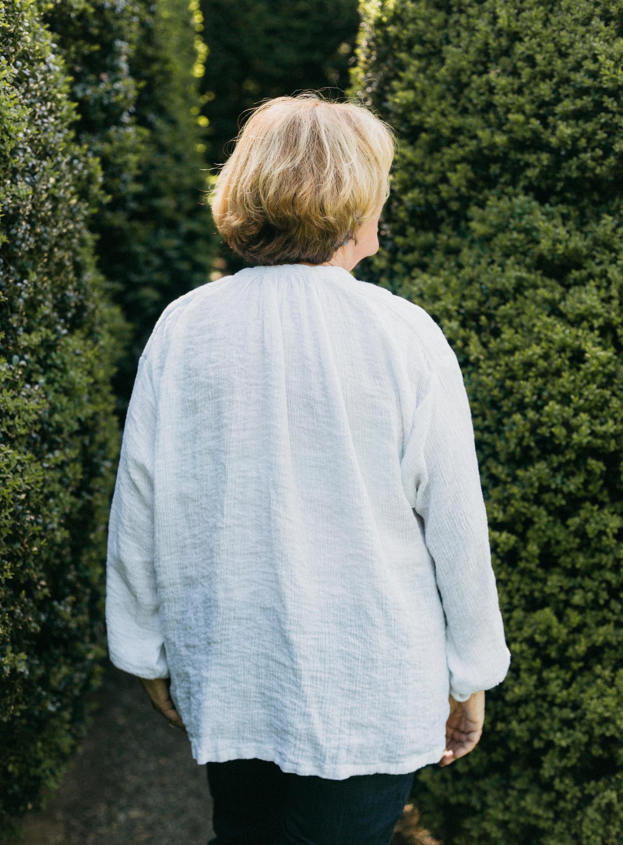 Older woman wearing French Cheesemaker's Smock made from white linen, standing by a large hedge with her back turned.