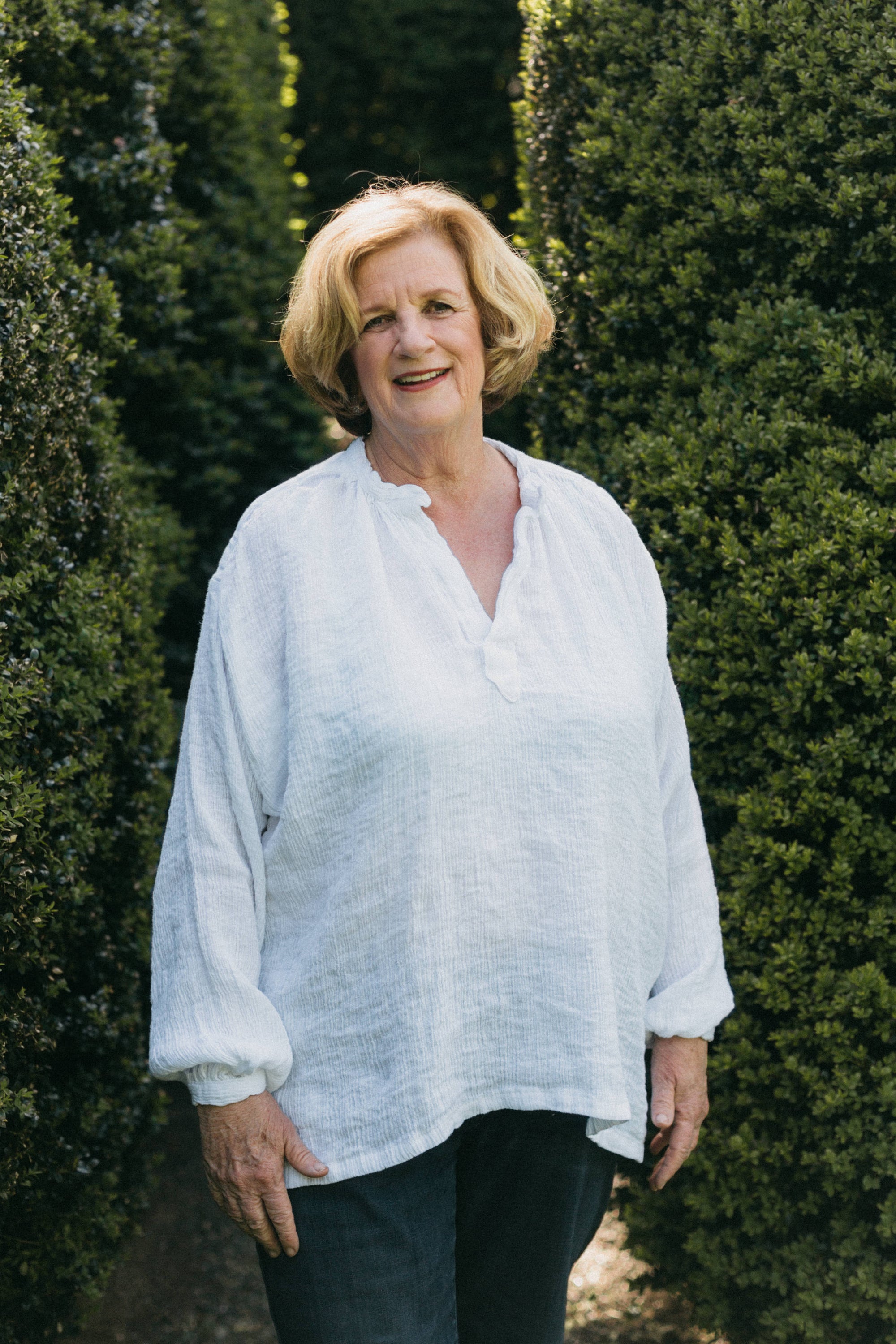 Older woman wearing French Cheesemaker's Smock made from white linen, standing by a large hedge