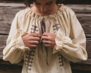 Picture of young woman adjusting ties of drawstring.  Front view of Romanian blouse with gathered cuffs and neckline.  Shirt is a natural gauze fabric with drawstring and embroidery applied in same color floss.