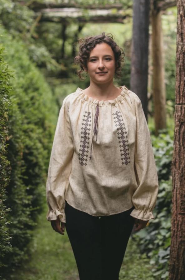 Young woman walking towards camera outdoors with greenery surrounding her. She is wearing 103 Romanian Blouse. Blouse is natural linen with traditional embroidery.  Paired with plain black pants.