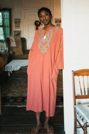 Young african american woman wearing long coral color version of 104 Egyptian shirt.  Shirt has grey applique around neck and on front yoke of top.  Model is leaning on door frame looking towards camera with hands in pockets.