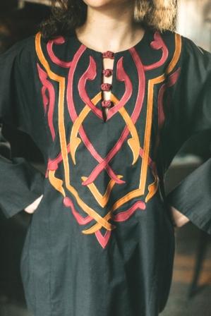 Close up photo of model in black Egyptian top.  Applique covers top from neckline to waist.  Four buttons are at closure of neckline slit.  Applique is two colors gold and coral.