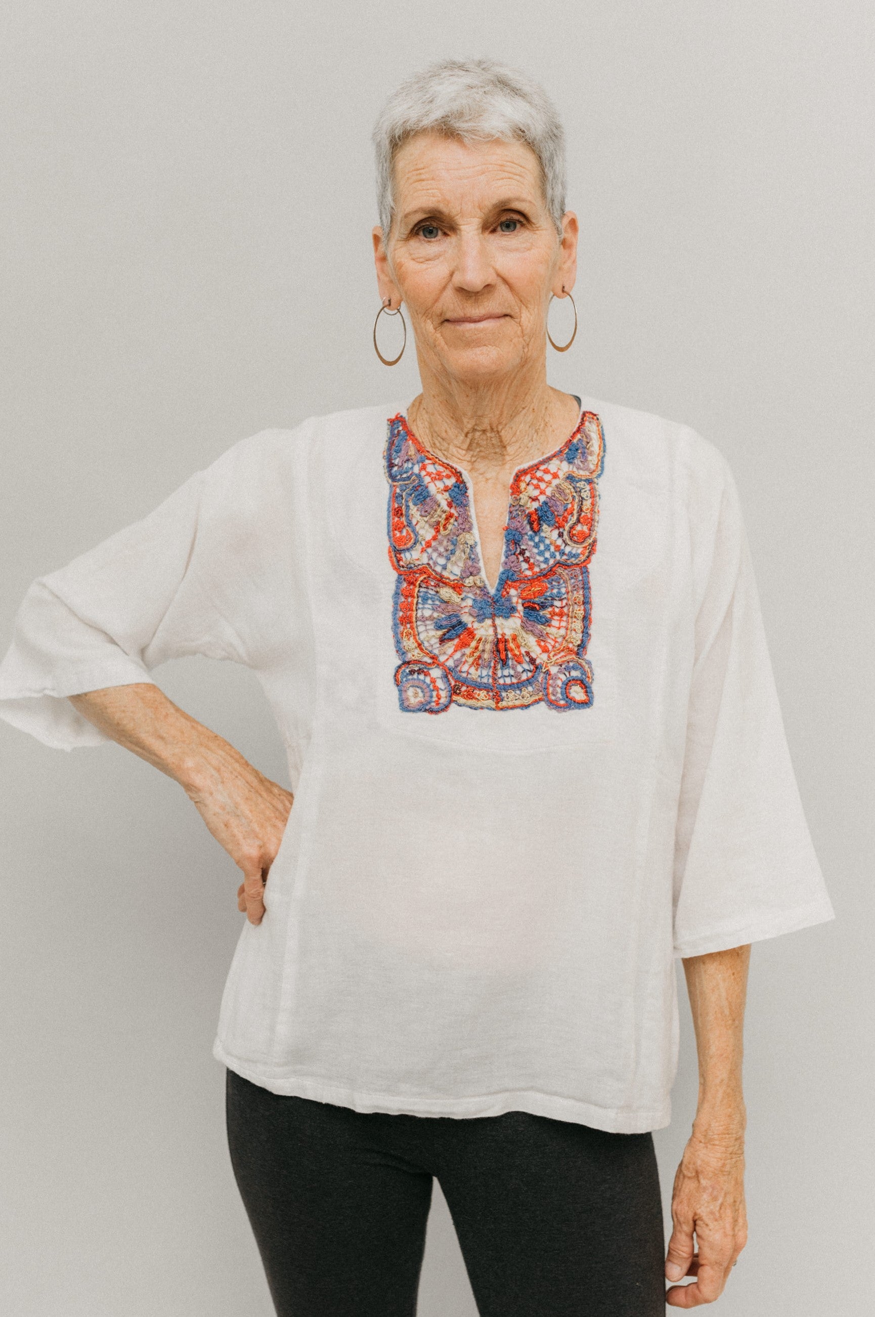 Older woman in short version of Egyptian top.  Top is made in a white gauze fabric with multi-colored embroidery on yoke.  Model has one hand on hip and poses with white backdrop.