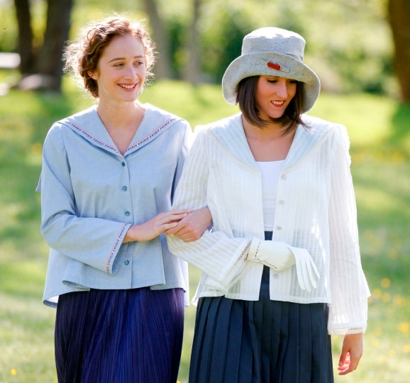 Two  women walking linking arms, surrounded by greenery. Woman on the left wearing #270 Metro Middy Blouse in blue and the woman on the right is wearing a hat with the Metro Middy Blouse in white.