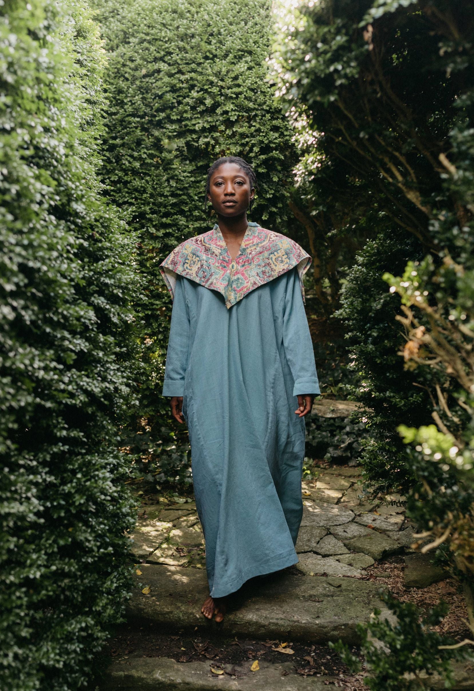 Black woman walking toward camera wearing a long blue dress with an embroidered triangular collar yoke.