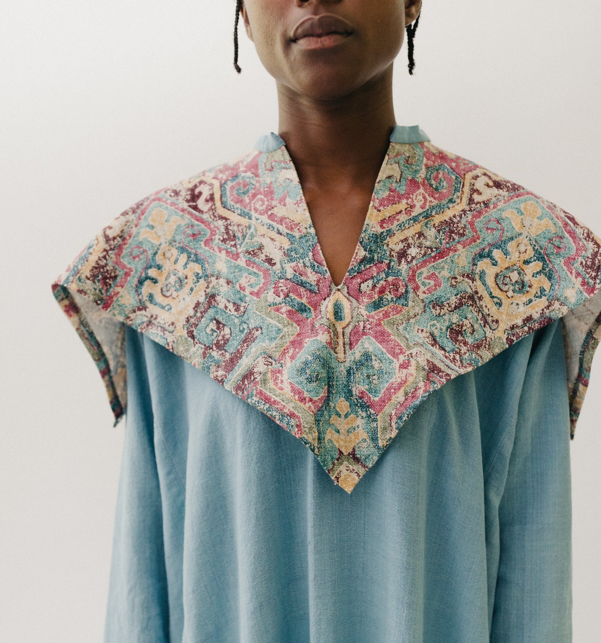 Black woman walking toward camera wearing a long blue dress with an embroidered triangular collar yoke. Close up on the collar with a white background.