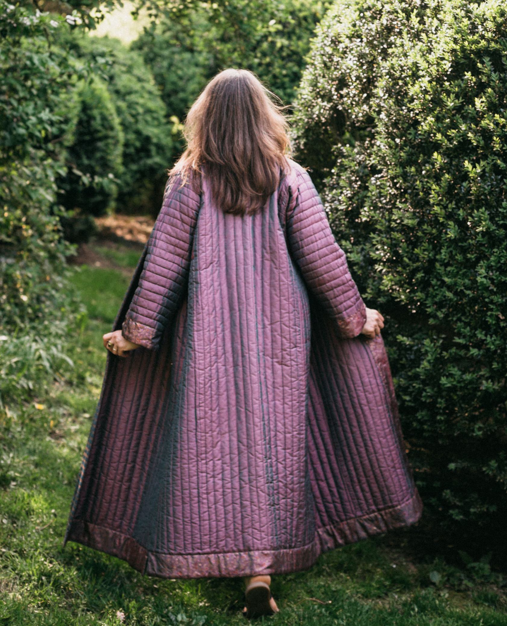 Back view of model walking away from camera outdoors wearing lavender silk Turkish coat.  Coat has full trapunto quilting and is ankle length. Model is holding jacket out at hip level showing full width.  