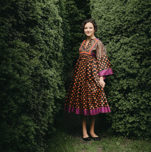 Young woman standing between two bushed with floral poppy print Afghan Nomad dress.  