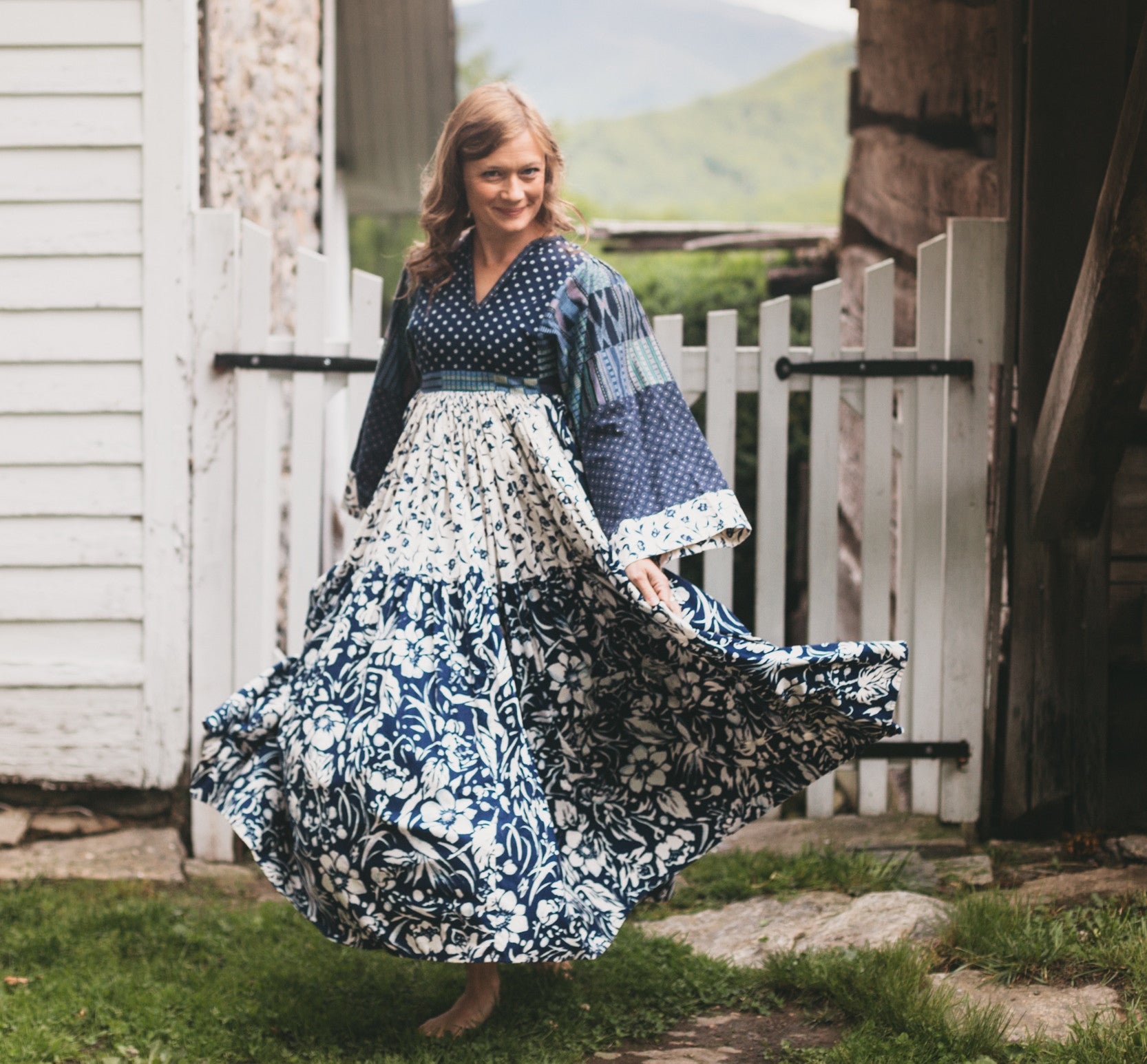 Woman walking towards camera in mulit fabric blue floral Afghan Nomad Dress.  Skirt is swishing as she walks.  White gate is behind her with a mountain view in distance