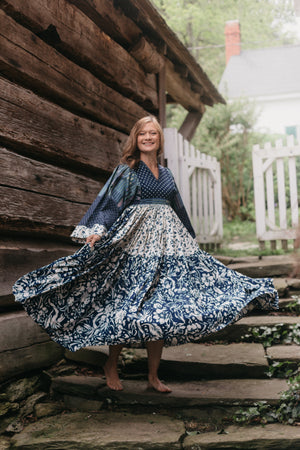 Woman twirling with full skirt in mid air.  Dress is multi fabrics all are various blue and white floral prints.  Woman is on stone stairs outdoors with wooden building behind her.