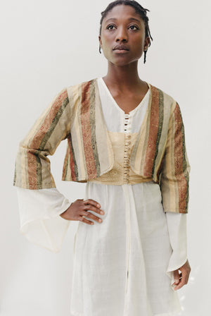 Black woman standing with hand on her hip in front of a white background.  She is wearing a white long sleeve entari, an embroidered gold vest, and a striped long sleeve jacket.