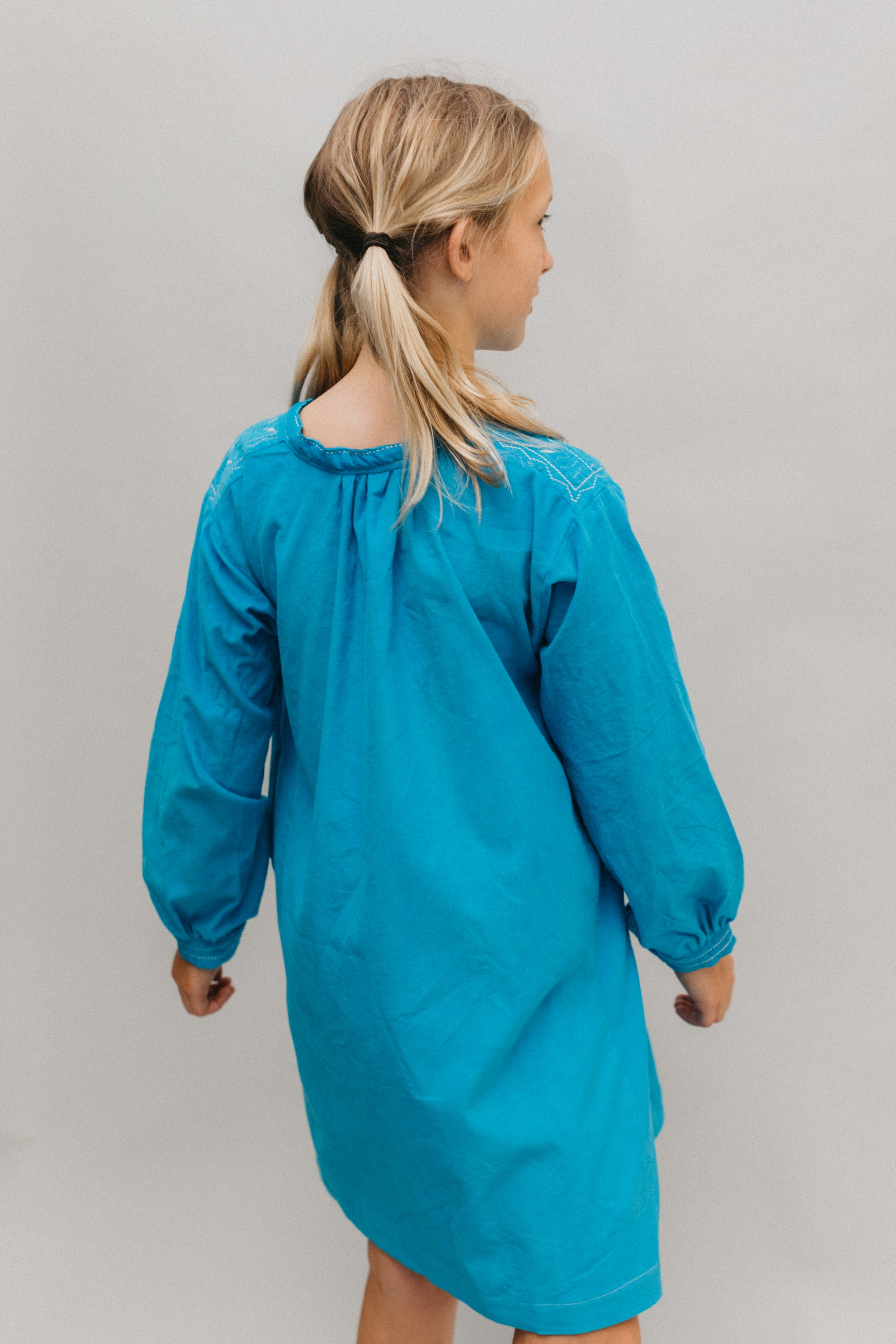 Photo of young girl wearing a blue corduroy Smock. Model had back to camera.  Photo shows gathering at neckline and back view.