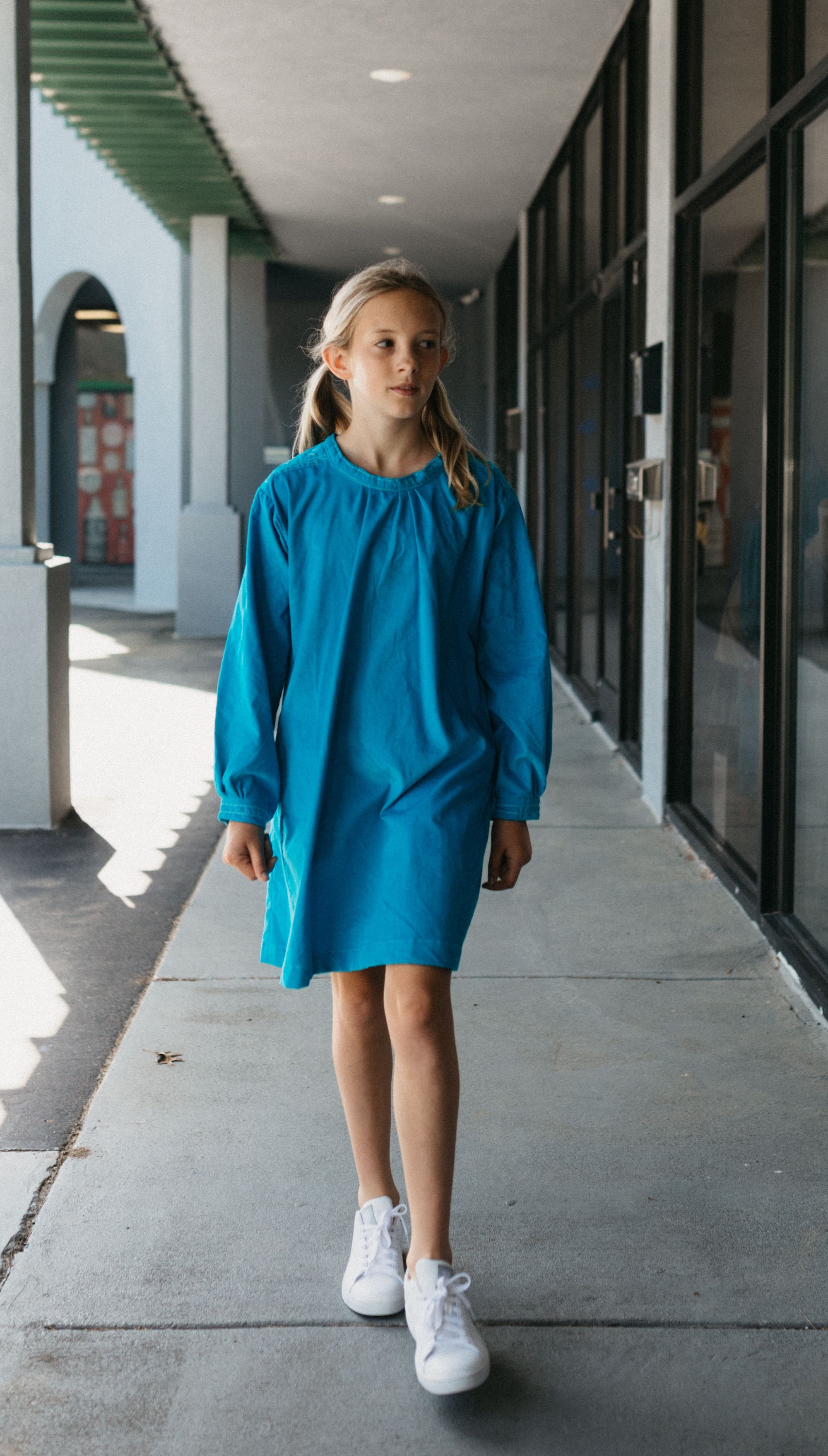 Photo of young girl wearing a blue corduroy Smock. Model is walking outside on sidewalk in front of stores. 