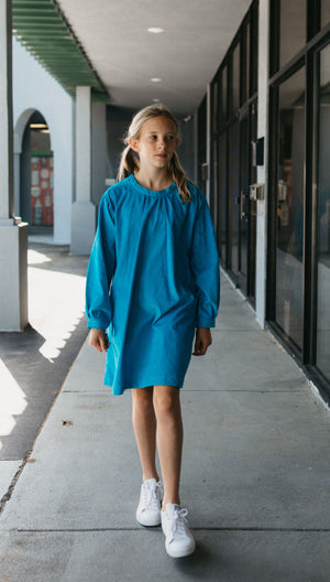 Photo of young girl wearing a blue corduroy Smock. Model is walking outside on sidewalk in front of stores. 