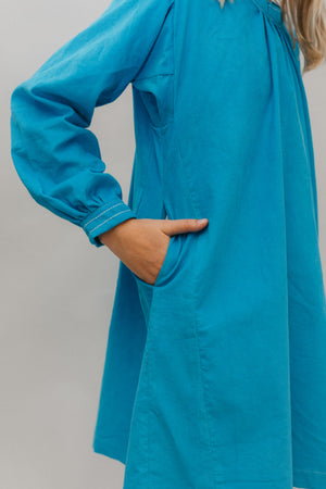 Photo of young girl wearing a blue corduroy Smock. Photo shows close up of sleeve cuff and side seam. Girl has her hand in the pocket at side.