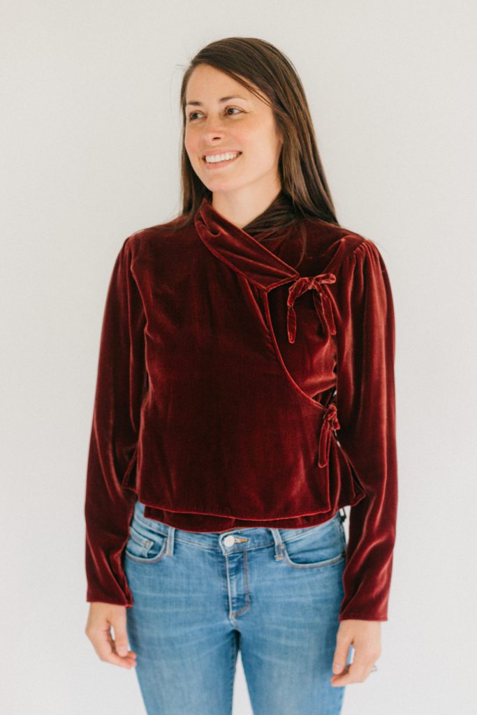 Woman in red velvet Nepali Top. Wearing jeans standing in front of white background.