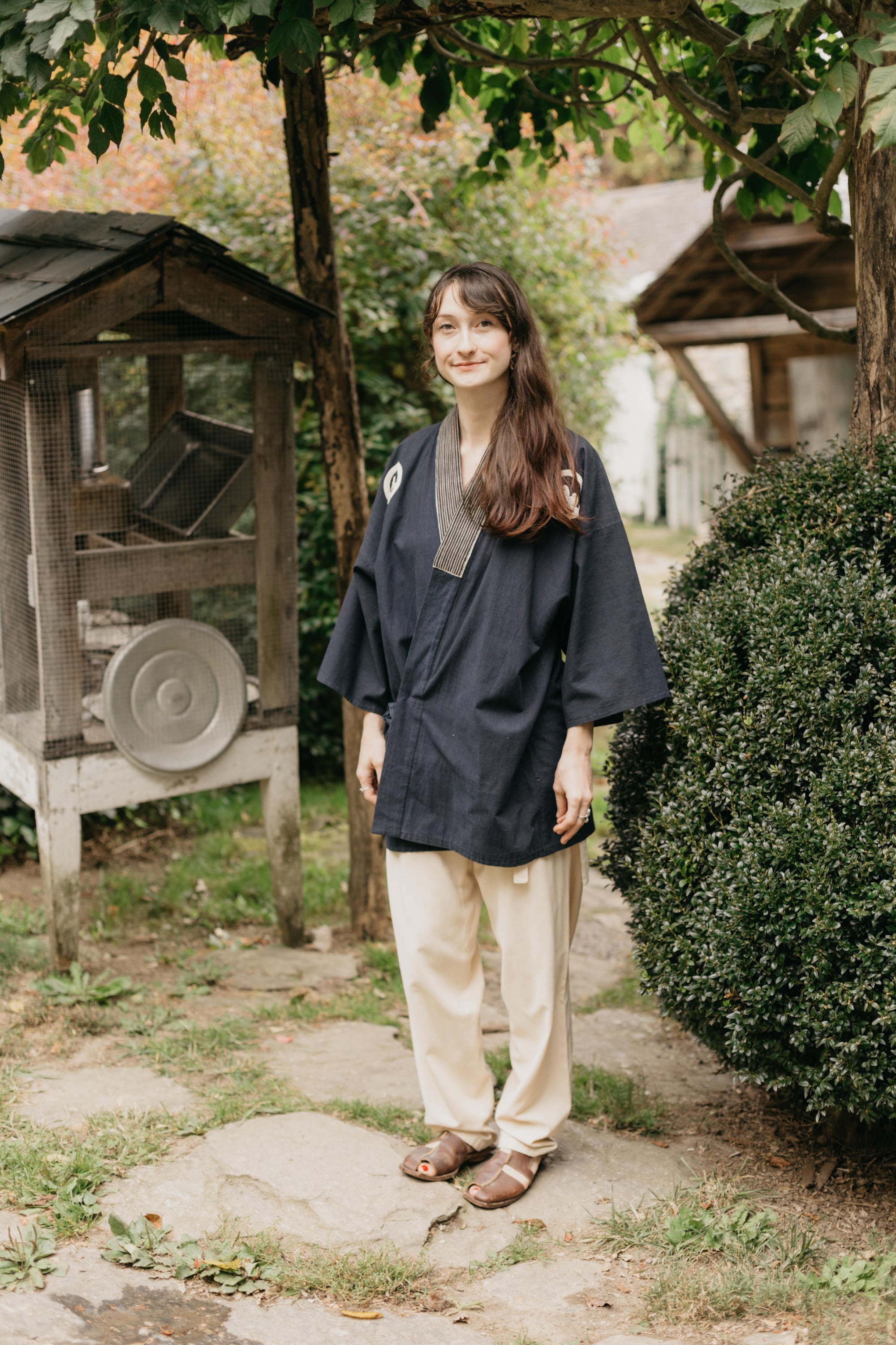 Japanese American woman wearing a navy blue Hippari and cream colored pants.  Standing outside by a shrub.