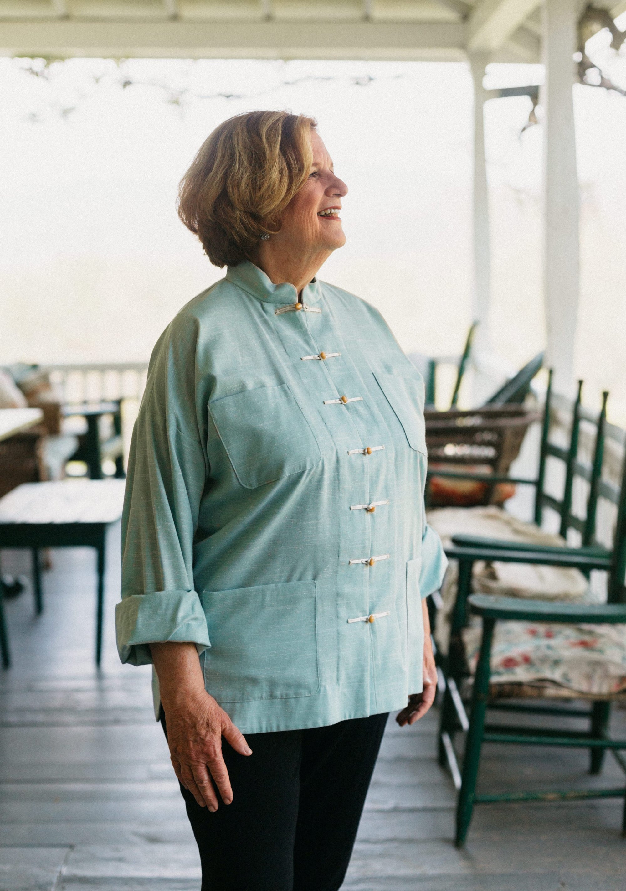 Older white woman smiling standing on the porch facing to the right, wearing Chinese pajama jacket in light teal.. 