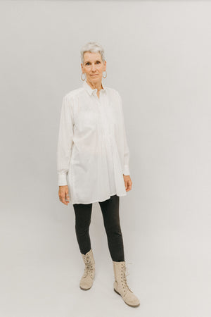 Woman standing in front of light grey background wearing a white Croatian shirt as a tunic with black leggings and boots.