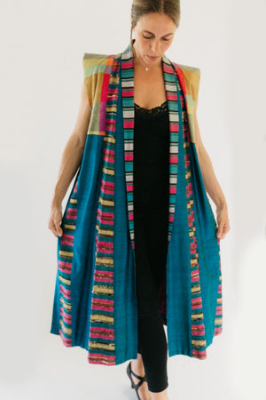 Woman wearing multi colored 118 Tibetan Panel coat.  Standing in front of white background holding sides out in hands.  coat is primarially blue with multicolored shoulders and trim.