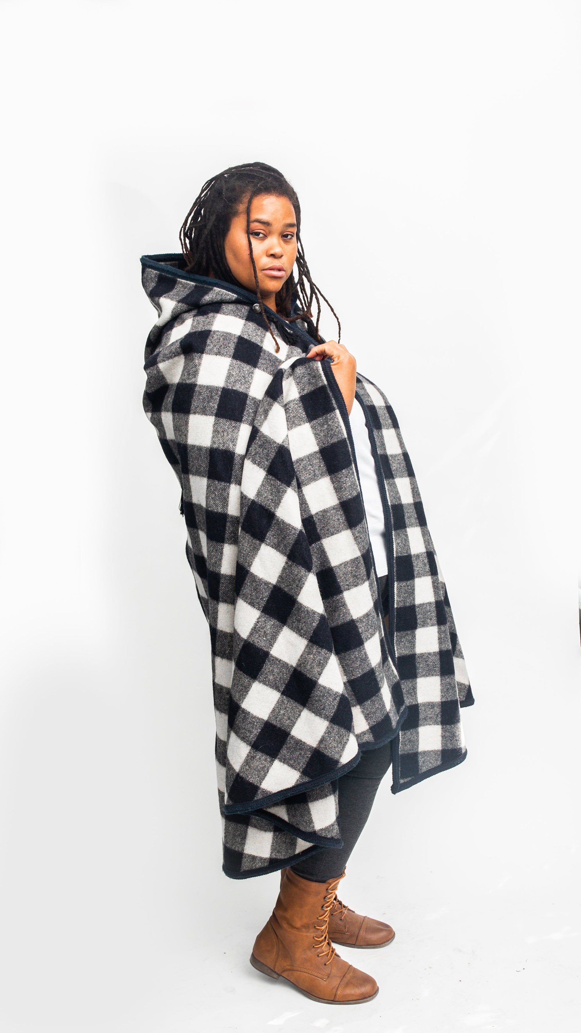 Black woman standing sideways in front of a white screen wearing a large black and white checked burnoose .