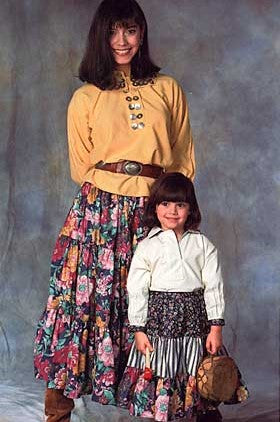 Pen and ink drawing by artist Gretchen Shields.  Woman is wearing 120 Navajo Blouse and skirt. Woman holds yarn and hand spiner in hands and stands in front of a loom.  Young girl holds a ball of yarn in hand.  A lamb is standing between the two.