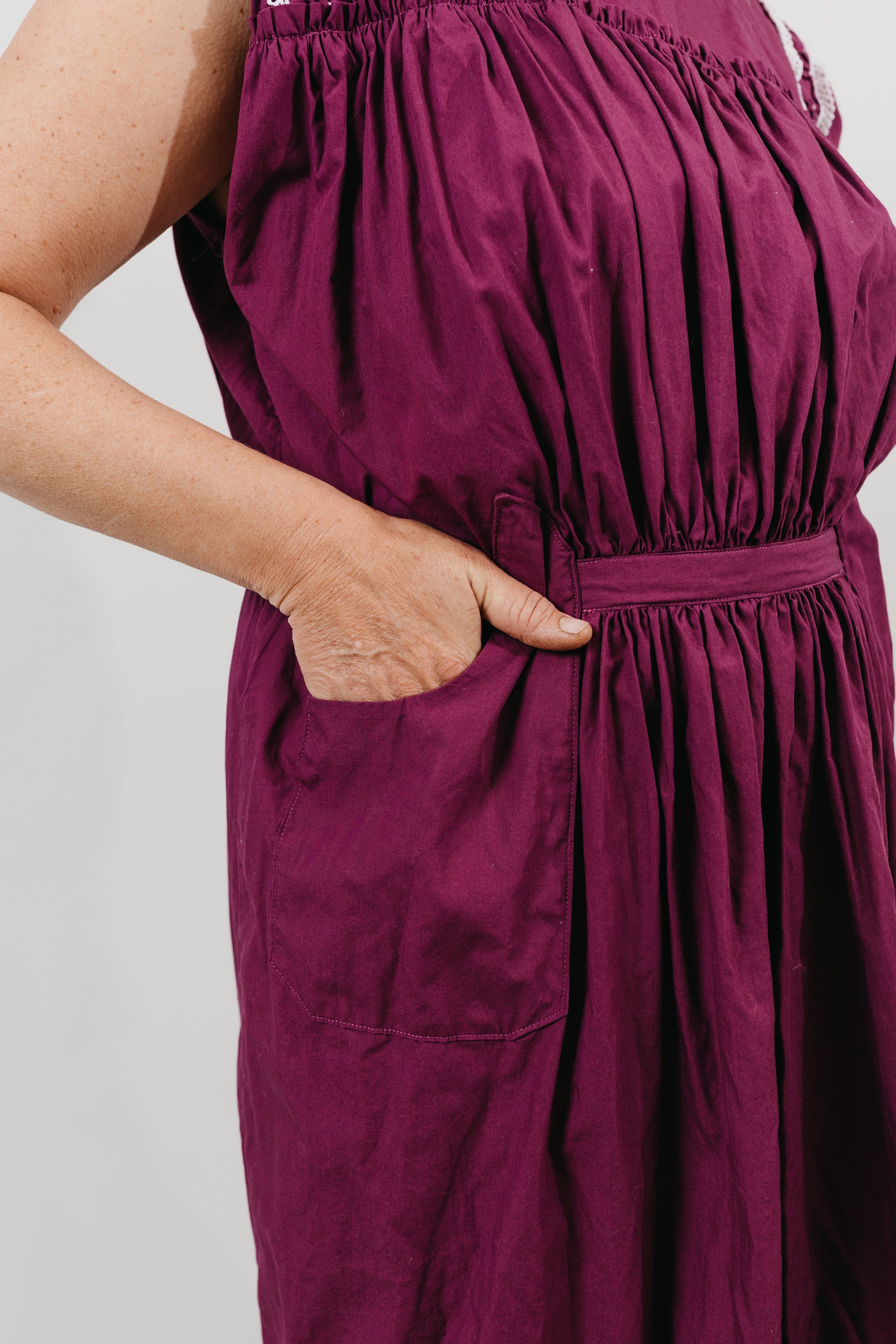 White woman wearing a purple sleeveless dress with embroidery at the shoulder yokes.  Standing in front of a white background.  Dress has pockets and is belted at the waist - close up of the pockets..