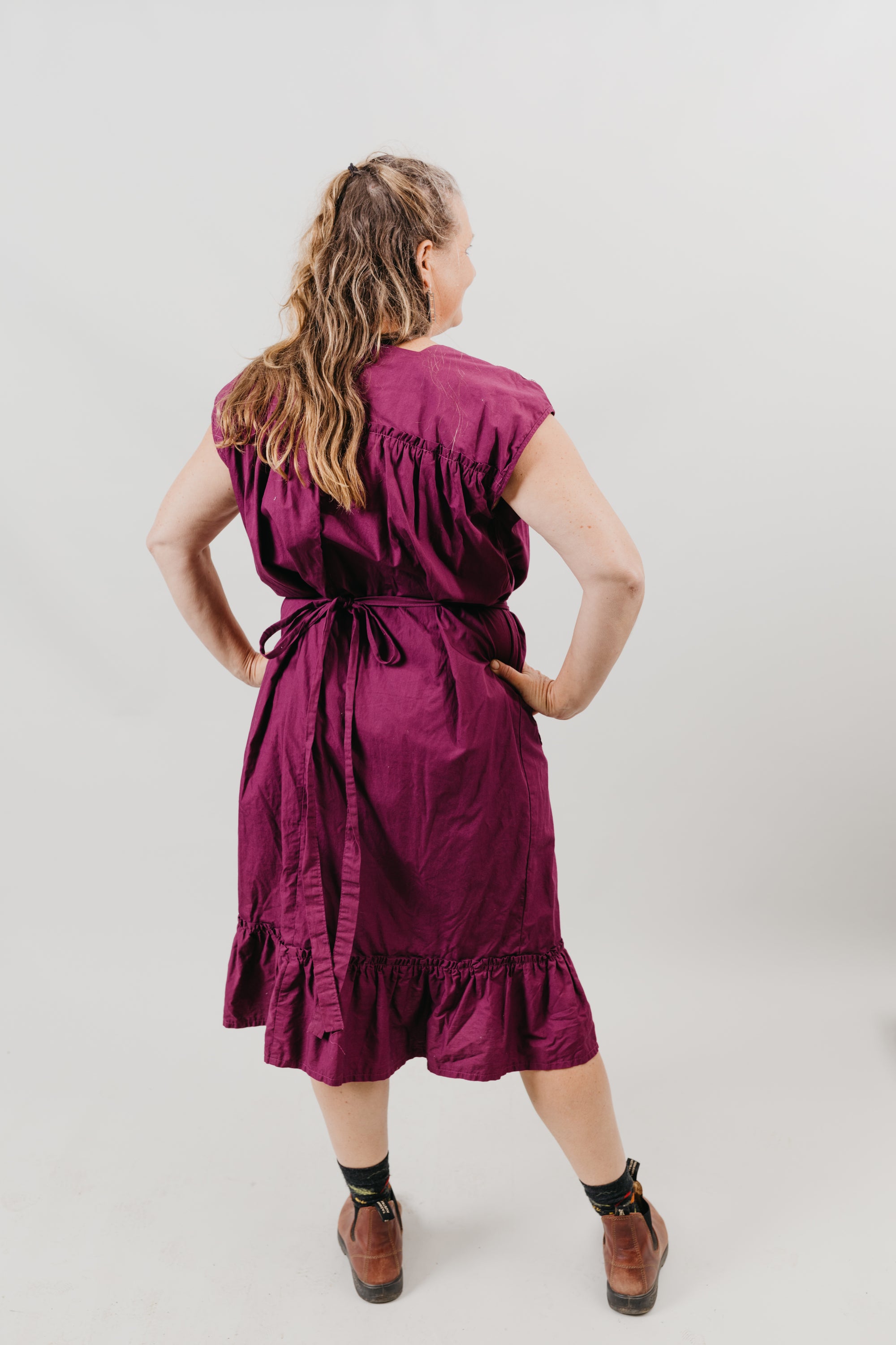 White woman wearing a purple sleeveless dress.  Standing with back to the camera in front of a white background.  Dress has pockets and is belted at the waist and has a flounce at the bottom.