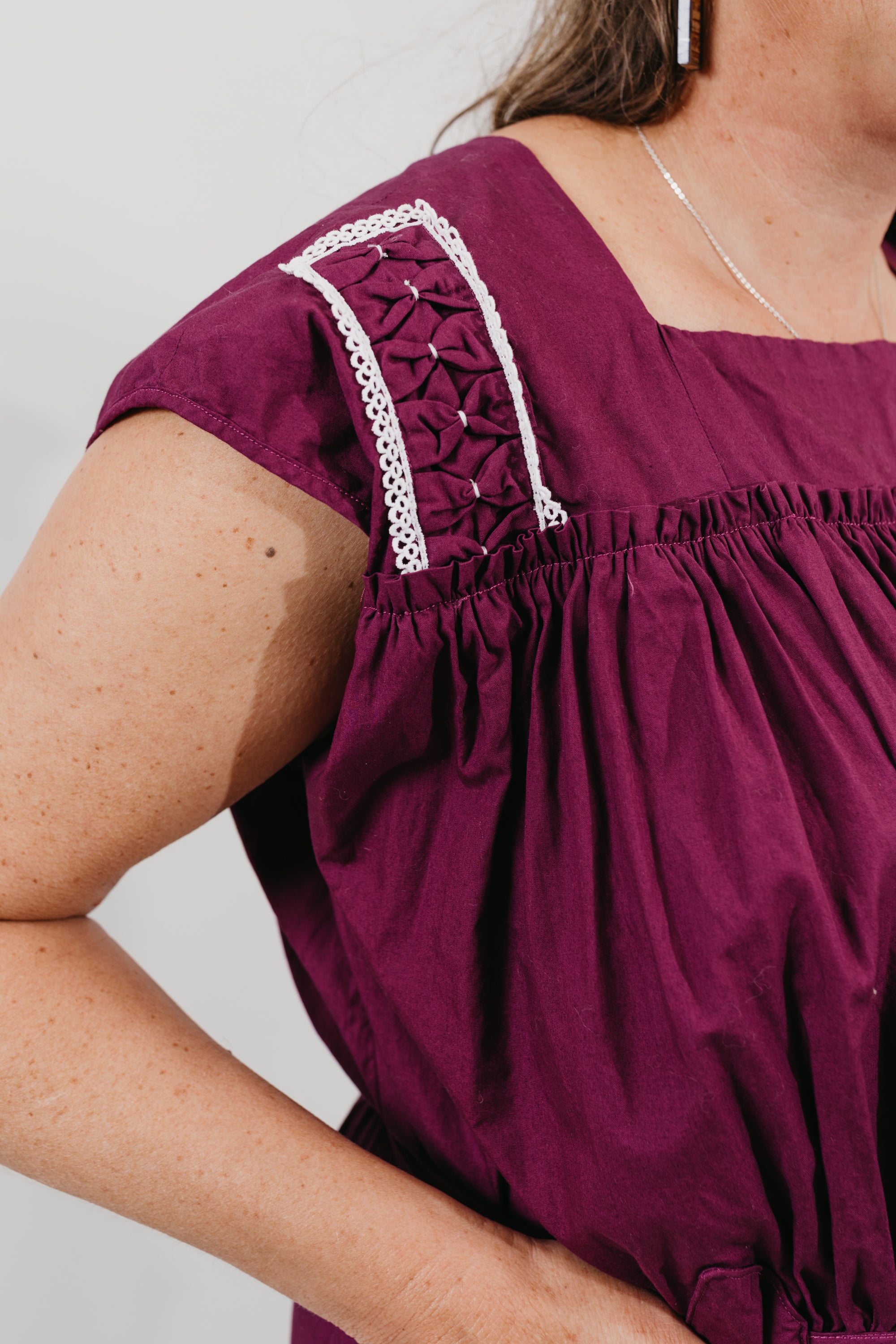 Close up of the shoulder embellishments - White woman wearing a purple sleeveless dress with embroidery at the shoulder yokes.  Standing in front of a white background.  
