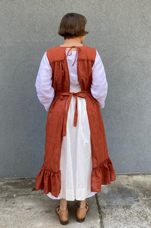 Back view of woman standing in front of a grey wall wearing a large sleeveless orange apron with pockets and a flounce at the bottom.