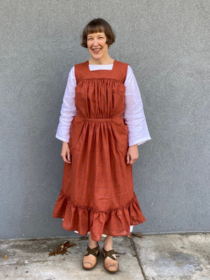 Woman standing in front of a grey wall wearing a large sleeveless orange apron with pockets and a flounce at the bottom.