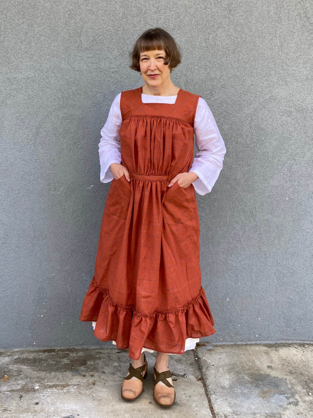 Woman standing in front of a grey wall wearing a large sleeveless orange apron with pockets and a flounce at the bottom.