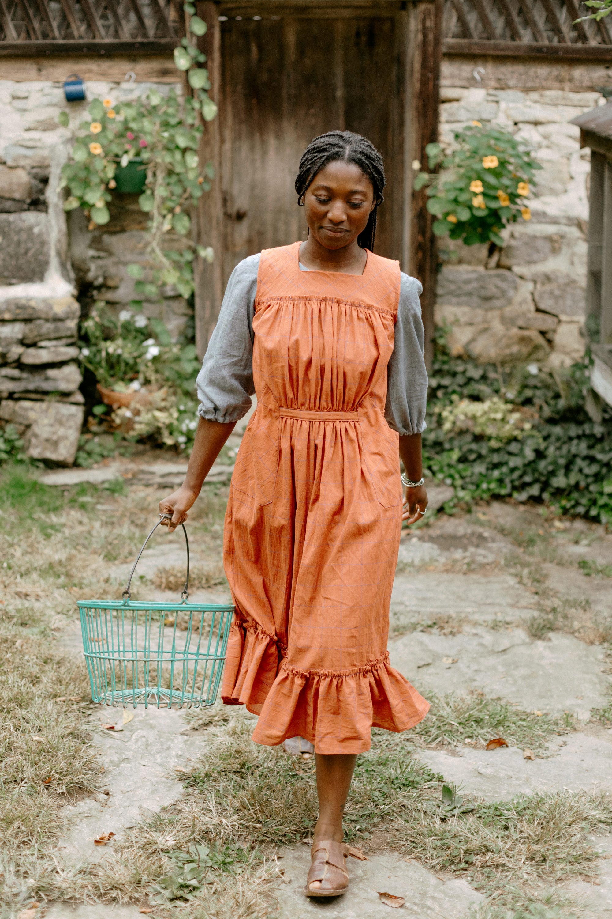 Black woman wearing a grey elbow-length sleeved Muumuu with an organic gabacha (apron) over the dress.  She is carrying an egg basket and apron has a flounce.