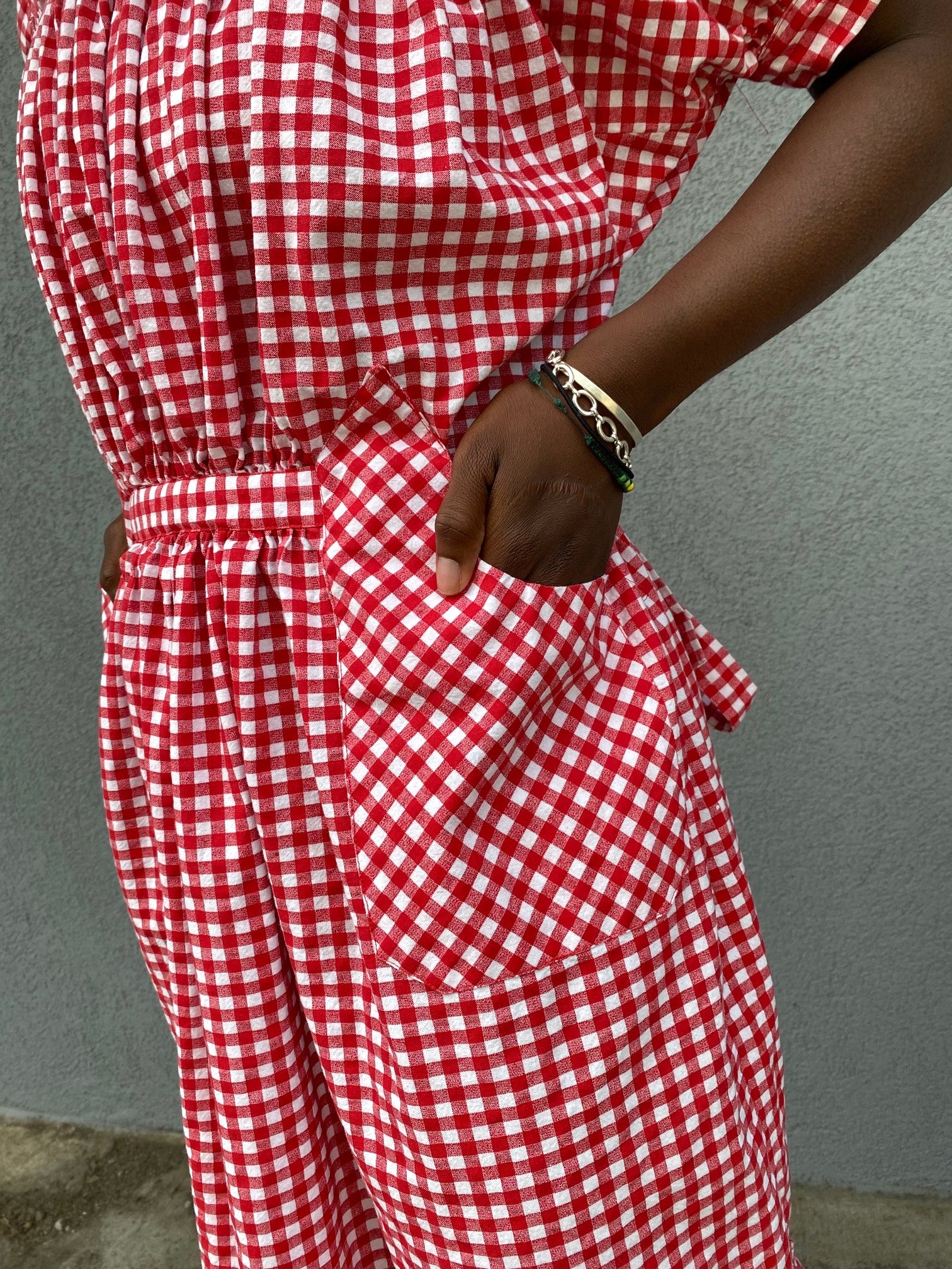 Close up of hand in a pocket of a woman standing in front of grey wall wearing a red and white check dress with short puffed sleeves and a bottom flounce.