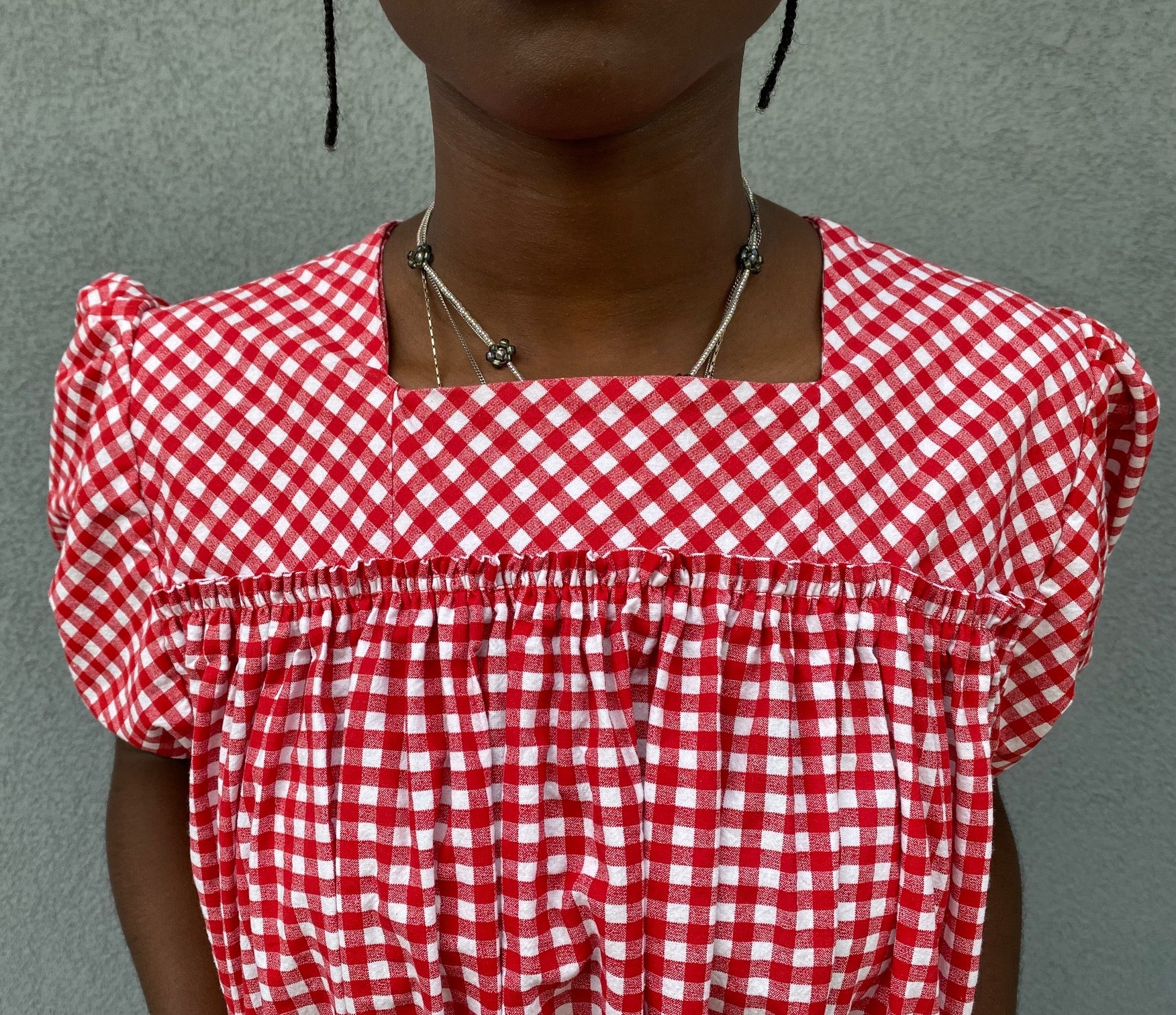 Close up of the bodice of Woman standing in front of grey wall wearing a red and white check dress with short puffed sleeves and a bottom flounce.