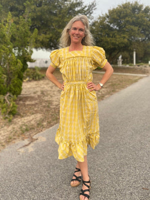 Woman wearing a yellow and white checked calf-length dress with puffed sleeves and pockets.  Standing outside in a street, wearing black strapped sandals.