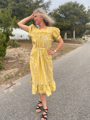 Woman wearing a yellow and white checked calf-length dress with puffed sleeves and pockets.  She is standing in a street with a hand by her head.  Dress has a flounce at the bottom