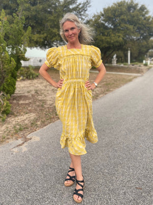 Woman wearing a yellow and white plaid calf-length dress, outside in a street. Dress has puffed sleeves and a flounce.