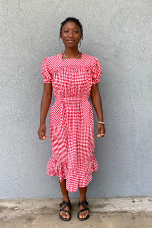 Woman standing in front of grey wall wearing a red and white check dress with short puffed sleeves and a bottom flounce.